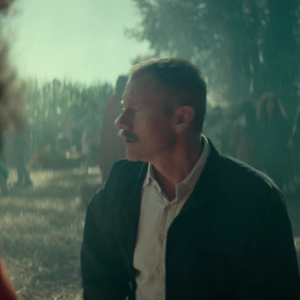 A mustached man in a dark jacket looks to his left outdoors in daylight in A24’s Slayground teaser.