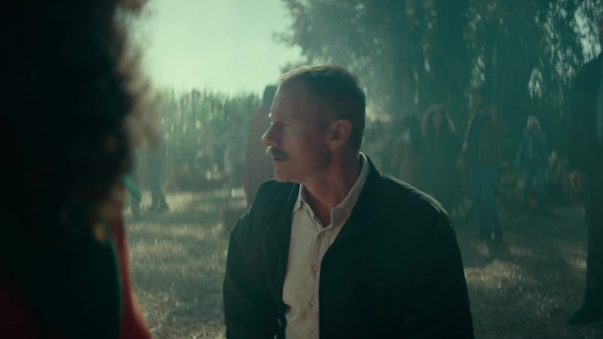 A mustached man in a dark jacket looks to his left outdoors in daylight in A24’s Slayground teaser.