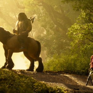 Brendan rides a horse through a sunlit forest as Prue stands beside her bicycle in Wildwood.