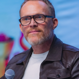 Paul Bettany wearing glasses and a dark jacket, speaking into a microphone at Los Angeles Comic Con 2025.