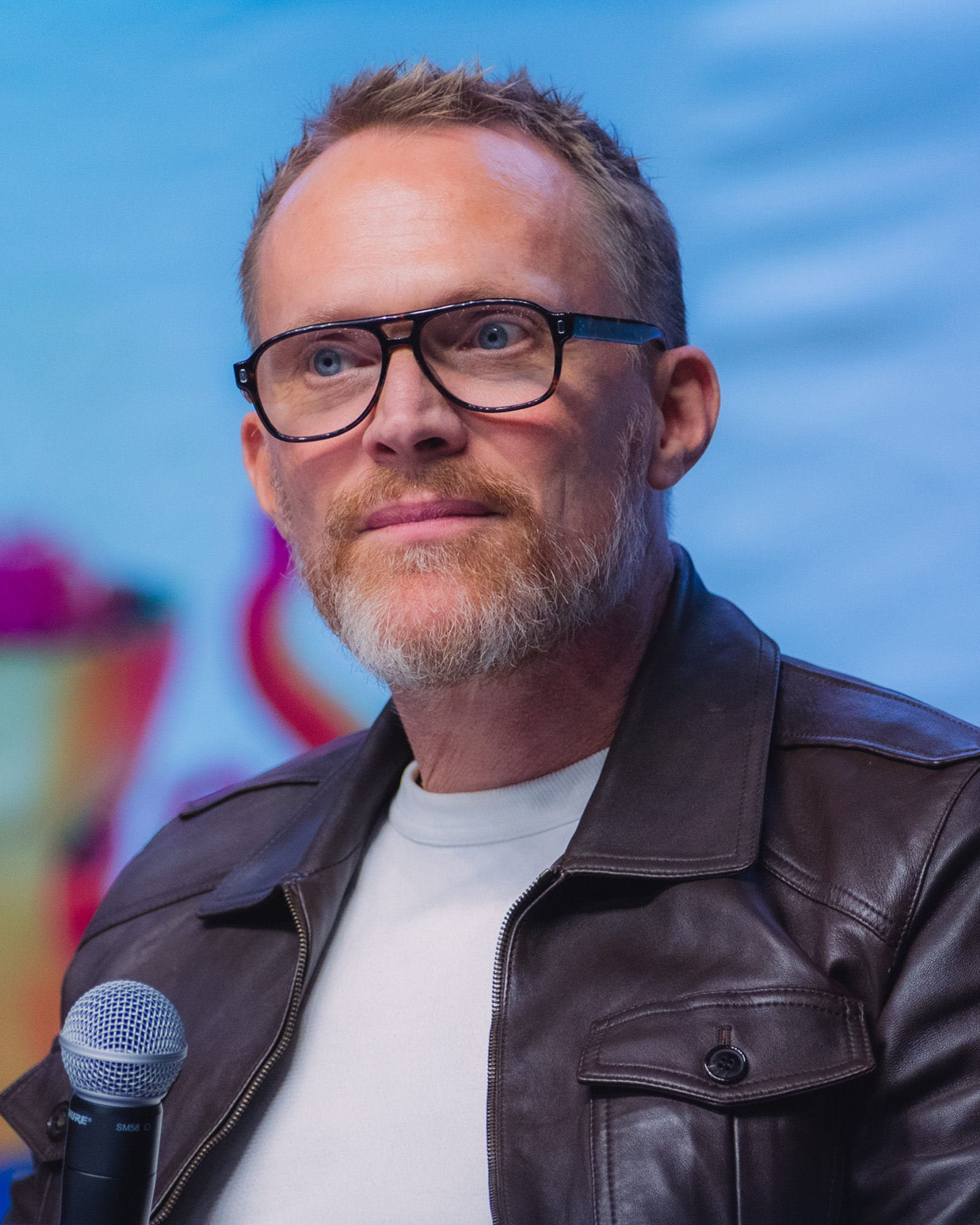 Paul Bettany wearing glasses and a dark jacket, speaking into a microphone at Los Angeles Comic Con 2025.